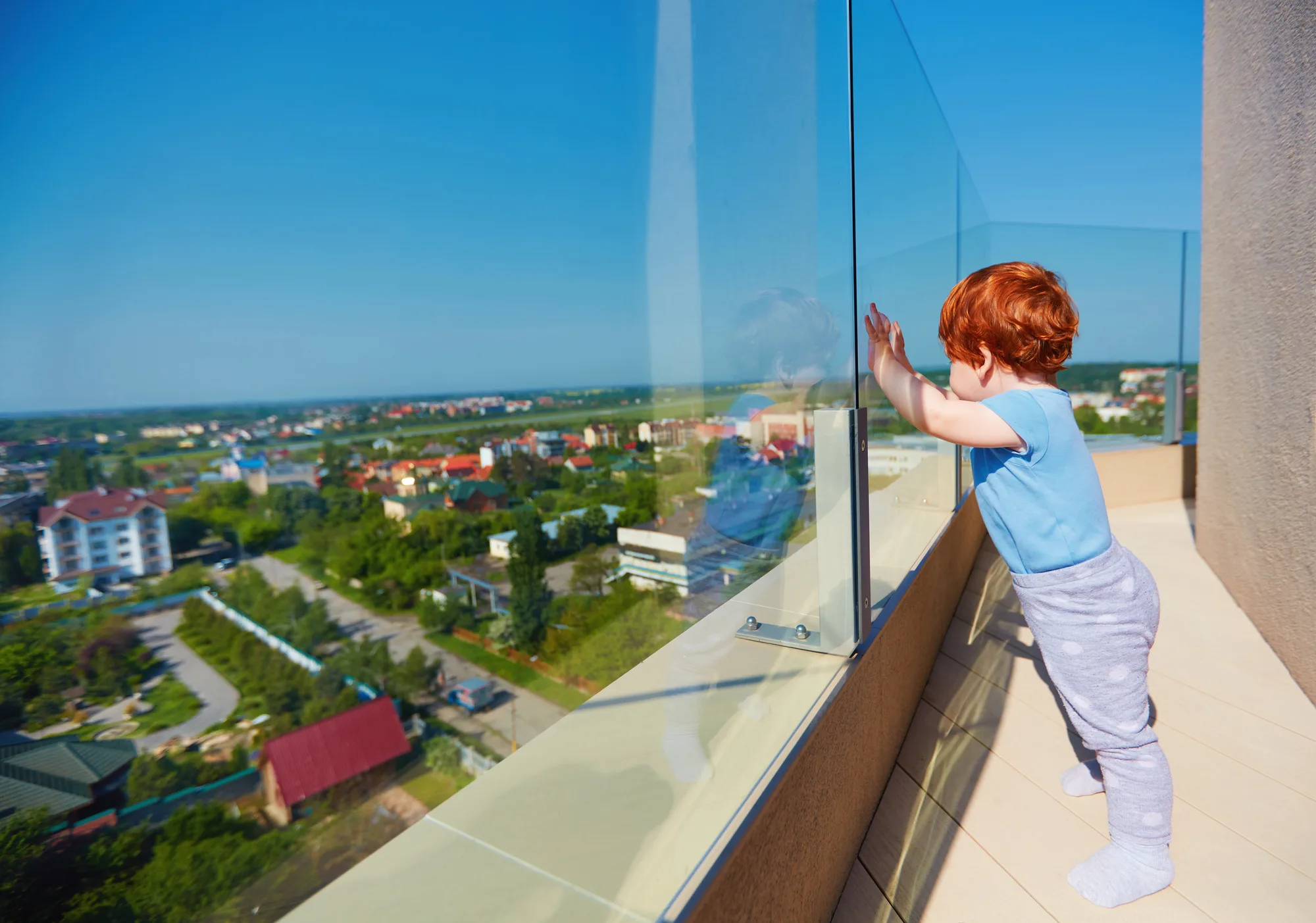 A young child reaching up to a frameless glass balustrade on a high-rise balcony
